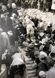 Widzimy grupę dzieci i dorosłych zgromadzonych wokół ceglanego muru w wykopie fundamentowym.Na pierwszym planie dzieci w szkolnych ubraniach (część w czapkach i płaszczach) przyklękają i przyglądają się pracy dorosłych.Kilku mężczyzn w garniturach i płaszczach bierze udział w symbolicznym murarskim akcie – może to być moment wmurowania aktu erekcyjnego.Zdjęcie dynamiczne, uchwycone z góry, pokazujące emocje – ciekawość, zaangażowanie.