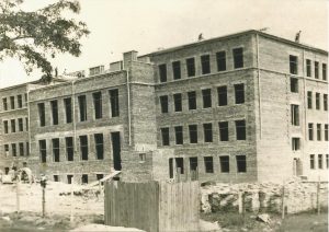 Przedstawia trzypiętrowy budynek szkoły wykonany z cegły, w stanie surowym, bez tynku i okien.Elewacja jest regularna, okna prostokątne rozmieszczone równomiernie w poziomach i pionach.Dach płaski, na którym widać kilku pracowników budowlanych.Teren wokół jest nieuporządkowany, z prowizorycznym ogrodzeniem z desek i kamiennym gruzem na pierwszym planie.Zdjęcie czarno-białe, wykonane prawdopodobnie latem.