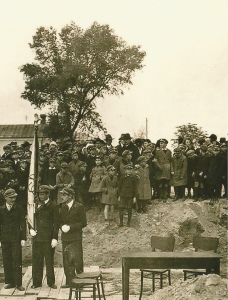 Ta sama scena co wyżej, ale pokazana bardziej z prawej strony.W tle tłum uczniów, nauczycieli i mieszkańców – dużo osób w czapkach i płaszczach, część dzieci ma dziecięce ubrania zimowe.Na pierwszym planie prosty stół i dwa krzesła, puste – najpewniej przygotowane do podpisania dokumentów lub wygłoszenia przemowy.