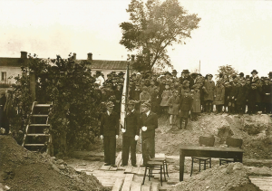 Trzech chłopców, prawdopodobnie uczniowie, w mundurkach szkolnych, trzymają flagę szkolną. Stoją na drewnianej platformie, w tle tłum dzieci i dorosłych – ceremonia patriotyczna lub założycielska.