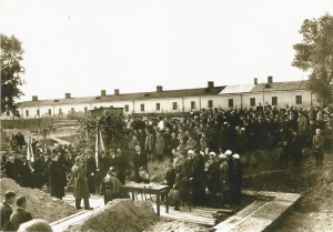 Bardzo duża grupa osób zgromadzona wokół drewnianej platformy, ustawionej nad wykopem budowlanym. Przy stole siedzą urzędnicy, trzymają dokumenty, obecny jest też poczty sztandarowe – uczniowie z flagą szkolną. Widoczny biały mur baraków lub budynków zaplecza budowy w tle.