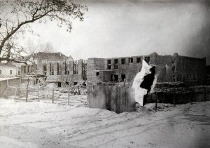 Budynek szkoły w stanie ruiny, prawdopodobnie po wojennych zniszczeniach. Brakuje dachu i części ścian, niektóre okna zamurowane lub wypalone. Śnieg pokrywa ziemię, w tle widoczne inne budynki, a na pierwszym planie zerwane betonowe konstrukcje.