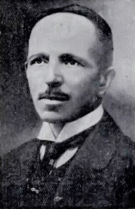 To czarno-białe zdjęcie portretowe przedstawia mężczyznę – tę samą osobę, co na poprzednim zdjęciu – w ujęciu popiersia, skierowaną nieco w prawo względem obiektywu. Tło jest jednolite i ciemne, dzięki czemu postać jest wyraźnie widoczna.Opis postaci:
Mężczyzna ma wysokie czoło, krótkie, ciemne włosy zaczesane do tyłu, a linia włosów wyraźnie cofnięta – sugerująca początki łysienia. Twarz pociągła, cera jasna, czoło i kości policzkowe dobrze widoczne. Ma regularne rysy twarzy, oczy wąskie, nos prosty i dość długi. Usta zamknięte, z cienkimi, ciemnymi wąsami nad górną wargą, które są przycięte starannie i symetrycznie. Mężczyzna nie ma brody.Na szyi widoczny jest wysoki kołnierz białej koszuli z charakterystycznymi, szeroko rozstawionymi końcami. Ma założony ciemny krawat lub muszkę oraz trzyczęściowy garnitur: marynarkę, kamizelkę i koszulę. Styl ubioru sugeruje, że zdjęcie zostało wykonane na początku XX wieku, prawdopodobnie w latach 1910–1930. Garnitur jest ciemny, najpewniej czarny lub granatowy, uszyty z gładkiego materiału.Wyraz twarzy i postawa:
Mężczyzna patrzy lekko w bok, z wyrazem skupienia i powagi. Twarz jest spokojna, ale zarysowane mięśnie żuchwy sugerują siłę charakteru. Postawa wyprostowana, świadcząca o pewności siebie i stanowczości.Cel zdjęcia:
Prawdopodobnie zdjęcie wykonane zostało do celów oficjalnych – np. do dokumentów, albumu, publikacji szkolnej lub państwowej. Portret ten ma klasyczny charakter urzędowy lub edukacyjny.Podsumowanie:
Zdjęcie przedstawia osobę z przełomu XIX i XX wieku, najpewniej nauczyciela lub urzędnika wyższego szczebla – z dużym prawdopodobieństwem jest to ponownie Wiktor Strzembosz, pierwszy dyrektor Gimnazjum Państwowego im. Staszica w Lublinie. Styl ubioru i charakter zdjęcia wskazują na osobę o wysokim statusie społecznym, cieszącą się szacunkiem.