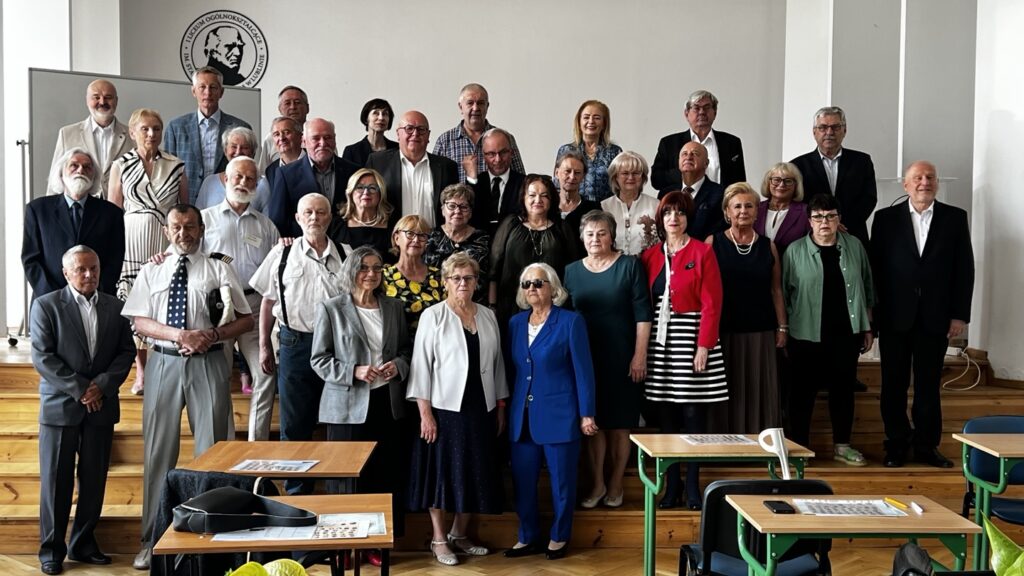 Rodzaj zdjęcia: Zdjęcie grupowe, zrobione wewnątrz sali lekcyjnej, przedstawiające kilkadziesiąt starszych osób. Prawdopodobnie są to absolwenci szkoły spotykający się po latach. Tło i otoczenie: Zdjęcie wykonano w dużej, jasnej klasie z białymi ścianami. Na tylnej ścianie, w lewym górnym rogu zdjęcia, znajduje się czarne godło lub logo szkoły. Logo zawiera wizerunek mężczyzny w profilu oraz napis wokół niego w języku polskim: „Zjazd Ogólnokrajowy Absolwentów” (część tekstu może być częściowo zasłonięta). W tle po lewej stronie – biała tablica. Pomieszczenie jest oświetlone światłem dziennym (brak widocznych lamp). Osoby stoją na kilku stopniach (podwyższeniach drewnianych) – są ustawione w czterech rzędach, co ułatwia widoczność każdej osoby. Na pierwszym planie, przed grupą, znajdują się ławki i krzesła szkolne. Na dwóch ławkach leżą przedmioty: czarna torba, okulary, kartka z nadrukiem. Osoby na zdjęciu: Grupa składa się z 38 osób: 19 kobiet i 19 mężczyzn. Większość z nich to osoby starsze (prawdopodobnie w wieku 60+). Wszyscy pozują do zdjęcia – patrzą w stronę obiektywu, przeważnie z uśmiechem lub neutralnym wyrazem twarzy. Ubrania są eleganckie, wizytowe – dominują garnitury, marynarki, sukienki, spódnice, koszule. Część osób ma okulary korekcyjne. Wiele kobiet ma fryzury krótkie, ułożone; część z nich jest siwowłosa lub blond. Mężczyźni głównie krótko ostrzyżeni lub z siwymi brodami. Jedna kobieta w pierwszym rzędzie pośrodku wyróżnia się – ubrana jest w intensywnie niebieski garnitur i ciemne okulary przeciwsłoneczne, ma siwe, rozpuszczone włosy – być może jest osobą niewidomą lub niedowidzącą. Niektórzy mężczyźni mają na sobie krawaty, inni są w rozpiętych koszulach – styl elegancki, ale różnorodny. W grupie panuje przyjazna, wspólnotowa atmosfera. Elementy dostępności i interpretacji: Zdjęcie wykonane na zakończenie spotkania lub podczas zjazdu absolwentów – klimat nostalgiczny i uroczysty. Kontekst (sala lekcyjna, tablica, godło szkoły) sugeruje powrót do szkoły po latach – symboliczne miejsce wspomnień. Osoby stoją blisko siebie, co podkreśla więź, znajomość i wspólną historię.