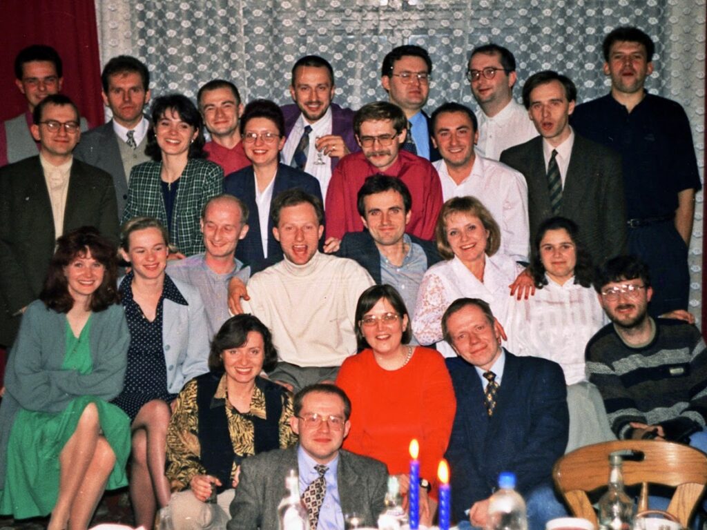 Grupa około 26 osób – kobiety i mężczyźni – pozuje w kilku rzędach we wnętrzu, prawdopodobnie podczas imprezy towarzyskiej. W tle firanka z koronkowym wzorem na oknie. Przed grupą stoi stół, na którym widoczne są butelki, szklanki, talerze i dwie niebieskie świece. Uczestnicy ubrani są elegancko, w marynarki, sukienki, koszule, niektórzy z kieliszkiem w ręku – atmosfera formalna, ale radosna. Zdjęcie ma lekko starodawny charakter – pochodzi prawdopodobnie z lat 90., co sugeruje styl ubioru i jakość fotografii.