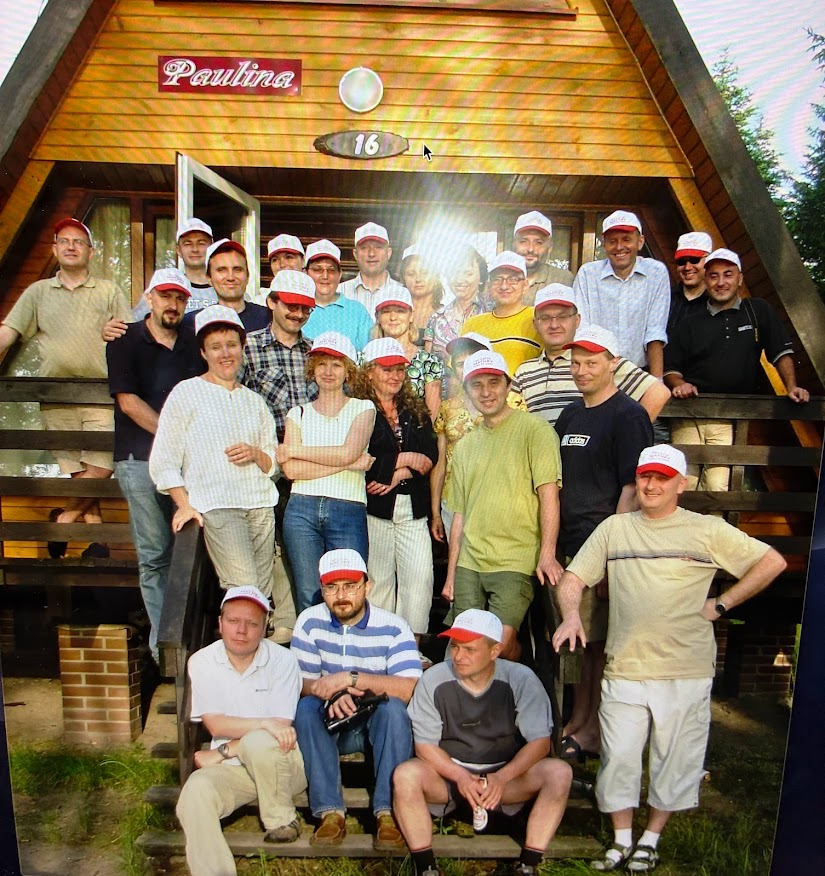 Około 30 osób pozuje na schodach i werandzie drewnianego domku letniskowego w kształcie litery "A". Nad drzwiami domek ma szyld z imieniem „Paulina” i numerem 16. Wszyscy uczestnicy mają na głowach białe czapki z czerwonym daszkiem, co sugeruje zorganizowane wydarzenie lub integrację. Grupa stoi na drewnianych schodach oraz werandzie. Osoby są ubrane swobodnie – koszulki, dżinsy, krótkie spodenki. W centrum zdjęcia – błysk światła od aparatu odbija się na twarzach części osób. W tle zieleń, drzewa iglaste.