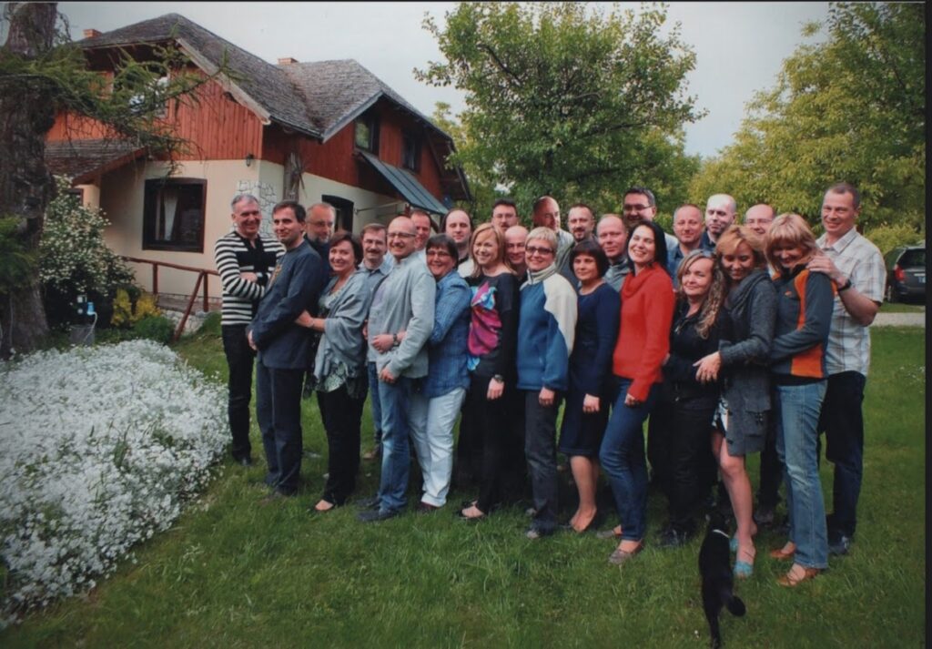 Na zdjęciu znajduje się około 30 osób stojących w dwóch rzędach, bardzo blisko siebie, wielu się przytula lub obejmuje. W tle widać dom z czerwoną i beżową elewacją oraz dachem z gontu. Na lewo od grupy – duże drzewo i biały krzew z kwiatami. W tle zieleń: drzewa i trawnik. Ubrania osób to głównie swetry, kurtki, bluzy – typowe dla wczesnej jesieni lub późnej wiosny. Z przodu, przy stopach grupy, widać czarnego kota z białą łatką, który kieruje się w stronę ludzi. Wszyscy uśmiechnięci, atmosfera przyjazna i ciepła.