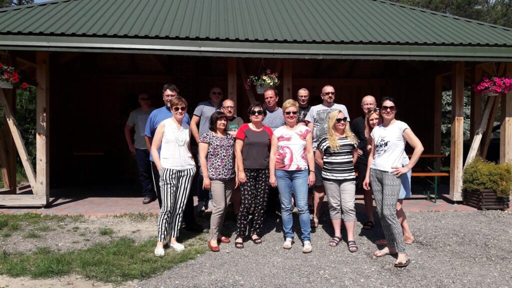 Zdjęcie przedstawia około 17 osób – kobiety i mężczyźni w różnym wieku – stojących przed dużą, drewnianą wiatą rekreacyjną z zielonym blaszanym dachem. Wiata znajduje się na utwardzonym placu z cegły klinkierowej, a przed nią lekko zakurzony teren z trawą i kamieniami. Po bokach widoczne są drewniane filary i wiszące kwietniki z różowymi kwiatami. Ludzie są ubrani letnio: t-shirty, bluzki, sandały. Jest słoneczny dzień, bardzo jasne światło, część osób mruży oczy. W tle, pod wiatą, widoczne drewniane ławki i stół piknikowy.