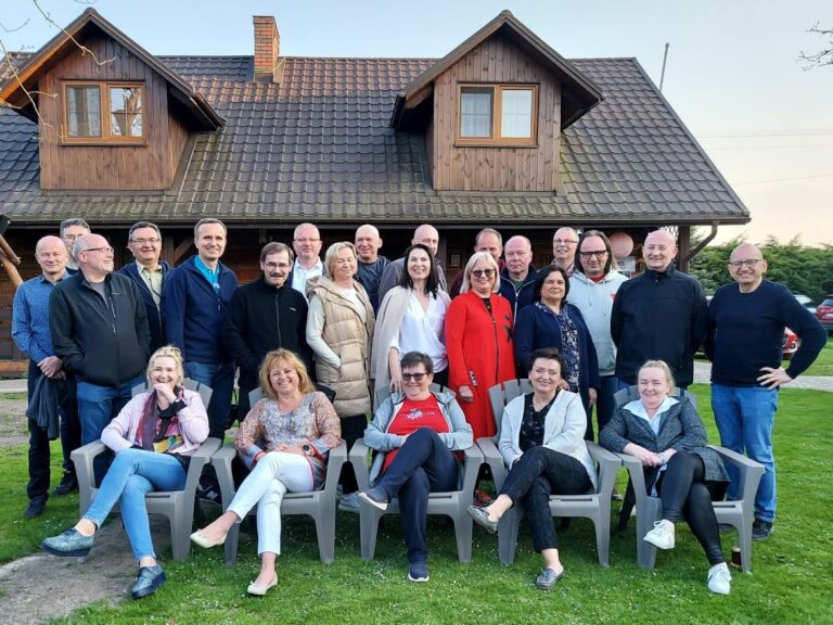 Grupa około 26 osób pozuje do wspólnego zdjęcia na tle dużego drewnianego domu w stylu wiejskim, z dachem krytym grafitową dachówką i dwoma małymi oknami dachowymi (lukarnami). Osoby stoją na trawie przed budynkiem – niektórzy stoją, inni siedzą na plastikowych fotelach ogrodowych w pierwszym rzędzie. Wszyscy uśmiechnięci, w różnym wieku, ubrani w casualowe, wiosenne stroje: swetry, kurtki, bluzy. Po lewej stronie domu widać huśtawkę ogrodową z drewna. Po prawej stronie, w tle, zaparkowane są samochody. Jest dzień – jasne niebo, miękkie światło, prawdopodobnie późne popołudnie.