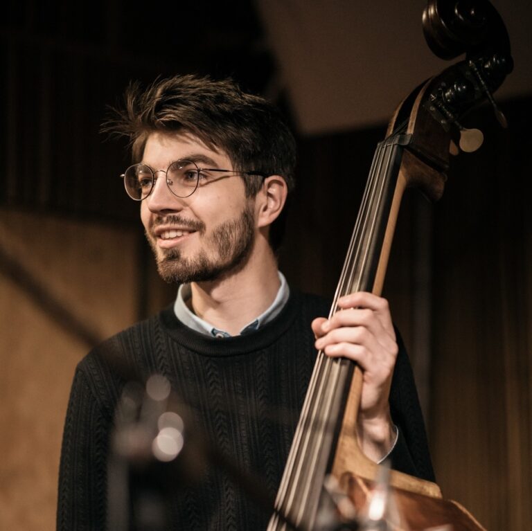 Naturalne, ciepłe zdjęcie mężczyzny grającego na kontrabasie. Zdjęcie prawdopodobnie wykonane podczas prób lub koncertu jazzowego. Opis szczegółowy: Młody mężczyzna z krótkimi, ciemnymi włosami i brodą. Ubrany w ciemny, dzianinowy sweter z fakturą w pionowe wzory i błękitną koszulę pod spodem. Na jego twarzy widoczny lekki uśmiech, głowa zwrócona lekko w lewo (z jego perspektywy), wzrok skierowany poza kadr. Na nosie ma okrągłe okulary w cienkiej metalowej oprawie. Prawą ręką trzyma gryf kontrabasu, który zajmuje prawą stronę kadru, a instrument wyraźnie dominuje w kadrze. Tło jest lekko rozmyte – widoczne drewniane elementy ściany, prawdopodobnie nagłośnieniowe lub akustyczne panele studia lub sali koncertowej. Atmosfera kadru jest ciepła i intymna, podkreślająca skupienie i pasję muzyka.