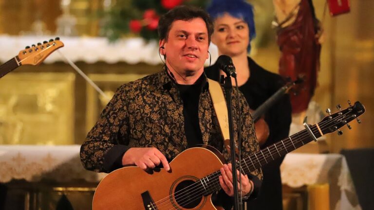 Zdjęcie przedstawia koncert lub próbę muzyczną w przestrzeni sakralnej – na tle ołtarza w jasnym, złocistym kolorze. W centrum kadru znajduje się uśmiechnięty mężczyzna, trzymający gitarę akustyczną. Ma na sobie czarną koszulę ze złotym, florystycznym wzorem, który nadaje elegancki charakter. Za nim, lekko w tle, stoi kobieta z niebieskimi włosami, uśmiechnięta, w czarnym stroju, trzymająca skrzypce. W tle widoczna jest dekoracja kościelna – białe obrusy, świąteczne kwiaty oraz figura świętego w czerwonym płaszczu. Scena oświetlona jest ciepłym światłem, co podkreśla kameralny i uroczysty charakter wydarzenia.