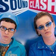 Dwie osoby stoją na tle niebiesko-czerwonej ściany z dużym napisem „Sound Clash”. Obie są wystylizowane w niebieskie stroje, utrzymane w spójnym, modnym stylu z elementami retro. Po lewej stronie stoi osoba w niebieskiej czapce z daszkiem, okrągłych okularach i błękitnej kurtce. Ma ciemne, prosto ścięte grzywki i poważny wyraz twarzy. Widać naszyjnik i kolorową koszulkę z grafiką. Po prawej stronie stoi osoba w jasnoniebieskich okularach przeciwsłonecznych, niebieskiej kurtce zapinanej na suwak i srebrnej biżuterii (kolczyki i naszyjnik). Ma blond włosy zaczesane do tyłu i również poważne spojrzenie. Oboje patrzą bezpośrednio w obiektyw, prezentując modowy wizerunek i spójny duet sceniczny. Tło i stylizacja sugerują udział w wydarzeniu muzycznym lub modowym.