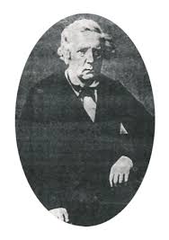 Zdjęcie przedstawia czarno-biały, owalnie przycięty portret starszego mężczyzny z XIX wieku. Zdjęcie ma charakter formalny i wygląda na wykonane w atelier fotograficznym.Wygląd mężczyzny:Mężczyzna ma siwe, falujące włosy, sięgające do wysokości uszu. Włosy są przedzielone z lewej strony jego głowy.Twarz jest pociągła, z wyraźnymi rysami: wysokim czołem, głęboko osadzonymi oczami, prostym nosem i lekko opuszczonymi kącikami ust. Wyraz twarzy jest poważny, wręcz surowy.Mężczyzna patrzy prosto w obiektyw, jednak jego wzrok wydaje się nieco zamyślony lub nieobecny.Ubrany jest elegancko, w ciemny surdut lub marynarkę, pod którą ma jasną koszulę i ciemną muchę lub kokardę zawiązaną pod szyją. Z kieszonki wystaje jasna chusteczka.Postawa i otoczenie:Mężczyzna siedzi, jego prawa ręka swobodnie opiera się na kolanie lub na poręczy krzesła. Dłoń jest częściowo widoczna – widać zgięte palce.Lewe ramię i dłoń są częściowo poza kadrem.Tło zdjęcia jest ciemne i jednolite – brak jakichkolwiek dodatkowych elementów scenografii. Zdjęcie skupia się wyłącznie na postaci.Całość ujęcia została umieszczona w owalnej ramce na tle prostokątnego obrazu, co nadaje fotografii klasyczny, archiwalny charakter.Charakter zdjęcia:
To typowy portret wykonany w stylu XIX-wiecznej fotografii – statyczny, poważny, przedstawiający osobę w pełnym stroju, bez uśmiechu. Może to być portret osoby historycznej lub naukowca, mający wartość dokumentalną.