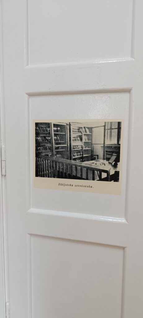 Zbliżenie białych drzwi. Na panelu drzwiowym przyklejona jest czarno-biała fotografia podpisana: „Biblioteka uczniowska”. Zdjęcie przedstawia wnętrze dawnej biblioteki – w tle znajdują się półki pełne książek, na pierwszym planie widać biurko z otwartą księgą, kałamarzem i piórem.