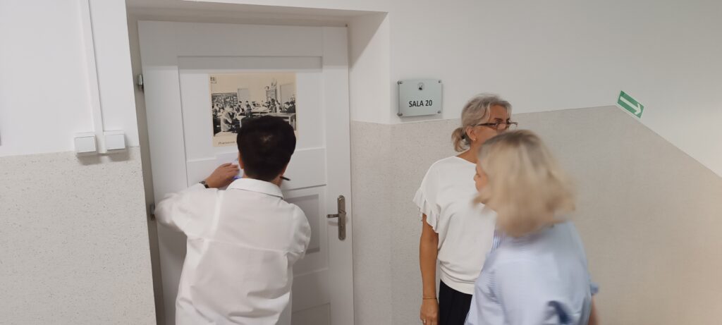 Przedstawia jasny szkolny korytarz. Na środku widać białe drzwi z metalową klamką po prawej stronie. Na drzwiach zawieszono czarno-białą fotografię z 1934 roku – przedstawia uczniów w pracowni biologicznej podczas lekcji przyrodniczej. Uczniowie siedzą przy długich stołach laboratoryjnych. Nad zdjęciem wisi mały obrazek. Na ścianie obok drzwi znajduje się tabliczka z napisem „SALA 20” umieszczona w srebrnej ramce. Obok drzwi stoją trzy osoby: jedna, zwrócona tyłem, coś przykleja do drzwi, a dwie pozostałe stoją obok i rozmawiają. Ściany są białe, dolna część korytarza wyłożona jest jasnoszarą lamperią. W prawym górnym rogu widać zielony znak ewakuacyjny ze strzałką w prawo.