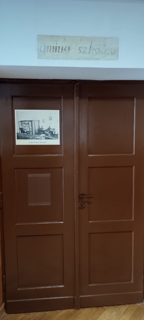 Podwójne, ciemnobrązowe, drewniane drzwi z metalową klamką po prawej stronie. Na jednym z paneli drzwi przyklejona jest czarno-biała fotografia podpisana: „Lokal Gminy Szkolnej”. Zdjęcie ukazuje wnętrze – kilka osób siedzi przy stołach, na ścianie wisi krzyż i portret. Obok stoi regał z otwartymi drzwiczkami, wypełniony książkami. Nad drzwiami na ścianie widnieje wyblakły napis „gmina szkoła”. Podłoga drewniana w jodełkę.