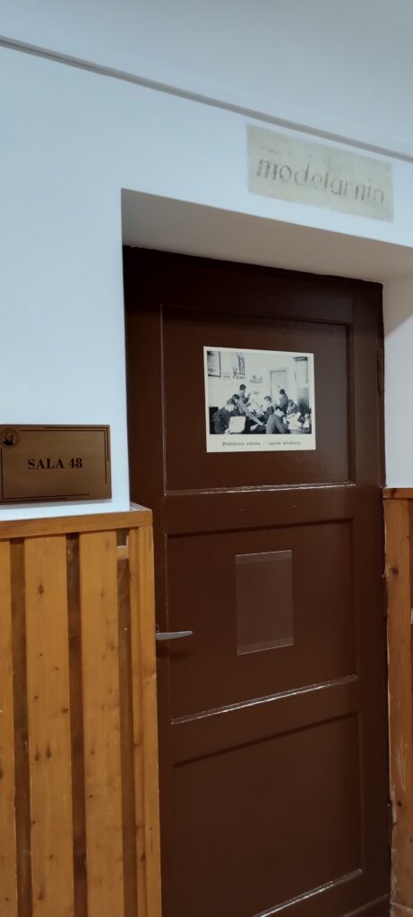 Ciemnobrązowe drzwi do sali z numerem 48. Tabliczka „SALA 48” znajduje się po lewej stronie drzwi, przy białej ścianie i drewnianej boazerii. Nad drzwiami widoczny wyblakły napis „modelarnia”. Na drzwiach przyklejona czarno-biała fotografia podpisana: „Modelarnia szkolna – zajęcia młodzieży”. Zdjęcie ukazuje grupę uczniów pochylonych nad stołami, pracujących nad modelami. W tle wiszą plakaty i rysunki techniczne.