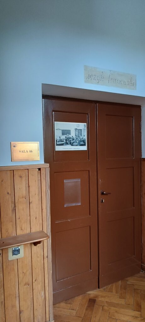 Ciemnobrązowe drzwi do sali z numerem 46. Po lewej stronie zamocowana złota tabliczka z napisem „SALA 46”. Nad drzwiami na ścianie widnieje wyblakły napis „język francuski”. Na drzwiach przyklejona czarno-biała fotografia podpisana: „Pracownia języka francuskiego”. Na zdjęciu uczniowie siedzą w sali lekcyjnej, w tle widać tablicę i nauczyciela prowadzącego zajęcia. Ściany wyłożone są drewnianą boazerią, a podłoga wykonana jest z drewnianych paneli w jodełkę.