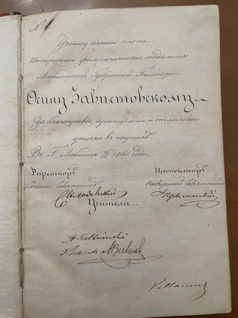 To wnętrze starej książki napisanej w języku rosyjskim, datowanej na 1860 rok.
Na górze strony widnieje numer „№ 1”, a poniżej kaligraficzny tekst zaczynający się od słów „Ученику пятого класса...” (czyli „Uczniowi piątej klasy…”).
Tekst zawiera imię i nazwisko ucznia – Zabłotnowskiemu – oraz informację, że został nagrodzony za dobre zachowanie i wyróżniające się wyniki w nauce.
Na dole widnieją podpisy dyrektora i nauczycieli, w tym wyraźny podpis Szwedowski i kilka innych odręcznych nazwisk w języku rosyjskim i polskim.
Papier jest pożółkły i ma drobne plamki starości.