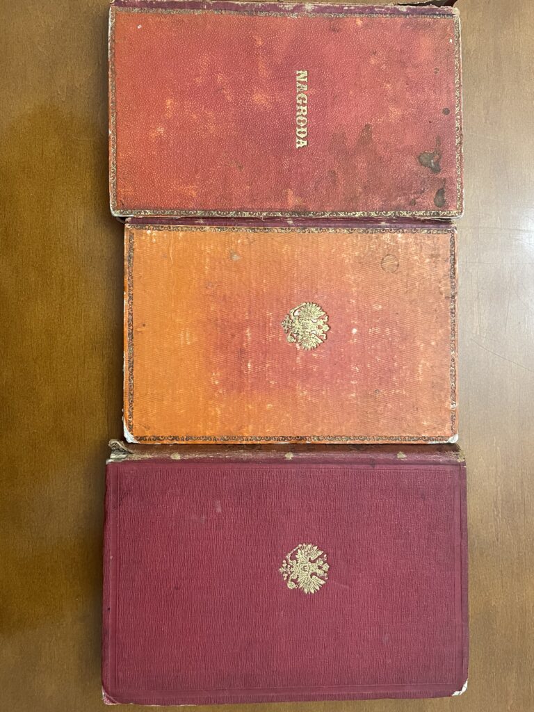 Na zdjęciu widać trzy stare książki ułożone pionowo, jedna nad drugą, na drewnianym stole w kolorze brązowym.
Od góry:Czerwona książka z napisem „NAGRODA” złotymi literami i ozdobną ramką – ta sama, co na poprzednim zdjęciu.Pomarańczowo-czerwona książka z delikatnym złotym ornamentem i złotym wytłoczonym orłem pośrodku.Ciemnoczerwona książka w nieco lepszym stanie, również ze złotym orłem pośrodku okładki.
Wszystkie książki mają twarde oprawy i widoczne ślady upływu czasu: otarcia, zagięcia, zadrapania i przetarcia na rogach.