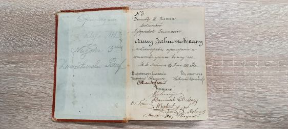 Na zdjęciu widać otwartą, starą książkę lub zeszyt leżący na jasnobrązowym blacie z drewnianą fakturą. Książka ma twardą oprawę w czerwonym kolorze z przetarciami na rogach. Strony są pożółkłe, co wskazuje na duży wiek dokumentu.
Na lewej stronie widoczne są ślady pisma ołówkiem – niewyraźne, prawdopodobnie podpis lub notatka.
Na prawej stronie znajduje się starannie wykaligrafowany tekst po rosyjsku, pisany czarnym atramentem ozdobnym pismem. U góry widnieje numer „№ 3”, poniżej kilka linijek tekstu, w którym wymienione jest nazwisko ucznia – Zabłotnowski (Zablotskij). Na dole strony widnieją podpisy kilku osób, prawdopodobnie nauczycieli i dyrektora gimnazjum, zapisane różnymi stylami pisma.