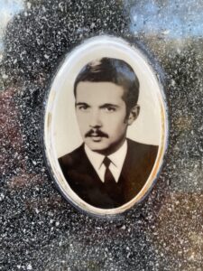 Na zdjęciu widoczna jest owalna, czarno-biała fotografia portretowa umieszczona na tle ciemnego, kamiennego materiału o ziarnistej, marmurowej fakturze. Zdjęcie znajduje się w owalnej, delikatnie wypukłej ramce o jasnym obramowaniu, prawdopodobnie porcelanowej, z drobnymi przebarwieniami i śladami czasu na krawędziach.Wewnątrz ramki przedstawiono mężczyznę w młodym wieku, prawdopodobnie z lat 60. lub 70. XX wieku, sfotografowanego od klatki piersiowej w górę. Ma krótkie, starannie zaczesane włosy, ciemne i gęste, przedziałek po lewej stronie. Twarz o regularnych rysach, jasnej karnacji, z wąsem i lekkim zarostem nad górną wargą. Oczy patrzą prosto w obiektyw, wyraz twarzy jest poważny, spokojny i skupiony.Mężczyzna ubrany jest elegancko: ma na sobie ciemny garnitur, białą koszulę z kołnierzykiem i ciemny krawat. Tło zdjęcia jest jasne i jednolite, bez dodatkowych elementów.Fotografia jest prawdopodobnie wtopiona w powierzchnię nagrobka – świadczy o tym kamienne, polerowane tło wokół niej, na którym widoczne są odbicia światła i drobinki pyłu. Całość sprawia wrażenie pamiątkowego portretu nagrobnego, zachowanego w dobrym stanie, choć z widocznymi śladami upływu czasu.