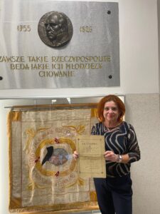 Kolorowa fotografia współczesna, wykonana wewnątrz budynku, najpewniej szkoły. Na pierwszym planie stoi kobieta w średnim wieku, ubrana elegancko w ciemne spodnie i bluzkę w ciemnogranatowe i złote wzory przypominające fale. Kobieta ma rude włosy i uśmiecha się, trzymając przed sobą wspomniane wcześniej stare świadectwo szkolne. Za nią znajduje się duży sztandar szkolny z haftowanym portretem mężczyzny w owalnej złotej ramie, otoczonej napisem „Ojczyźnie – Obywatelowi – S. Staszic”. Kolory sztandaru to złamany biały, złoty, pomarańczowy i różowy. Po lewej stronie zdjęcia widać fragment kamiennej tablicy pamiątkowej z metalowym medalionem przedstawiającym twarz mężczyzny z profilu. Obok napis w złotych literach: „TAKIE RZECZYPOSPOLITE, BĘDĄ JAKIE ICH MŁODZIEŻY WYCHOWANIE” oraz daty „1826” i „1755”. Tło jasne, wnętrze nowoczesne, z białymi ścianami i szklanymi gablotami.