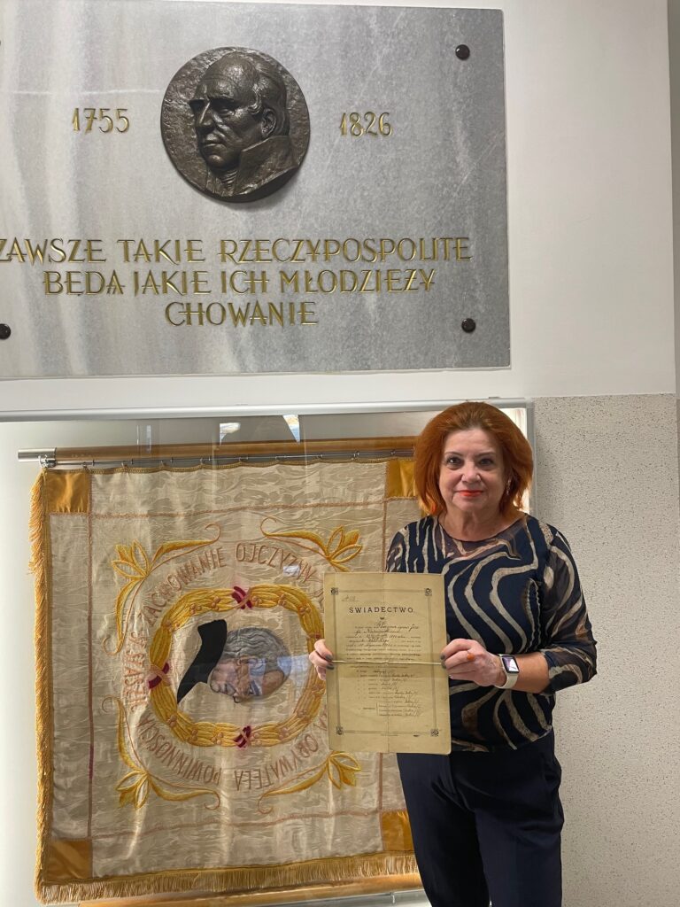 Kolorowa fotografia współczesna, wykonana wewnątrz budynku, najpewniej szkoły. Na pierwszym planie stoi kobieta w średnim wieku, ubrana elegancko w ciemne spodnie i bluzkę w ciemnogranatowe i złote wzory przypominające fale. Kobieta ma rude włosy i uśmiecha się, trzymając przed sobą wspomniane wcześniej stare świadectwo szkolne. Za nią znajduje się duży sztandar szkolny z haftowanym portretem mężczyzny w owalnej złotej ramie, otoczonej napisem „Ojczyźnie – Obywatelowi – S. Staszic”. Kolory sztandaru to złamany biały, złoty, pomarańczowy i różowy. Po lewej stronie zdjęcia widać fragment kamiennej tablicy pamiątkowej z metalowym medalionem przedstawiającym twarz mężczyzny z profilu. Obok napis w złotych literach: „TAKIE RZECZYPOSPOLITE, BĘDĄ JAKIE ICH MŁODZIEŻY WYCHOWANIE” oraz daty „1826” i „1755”. Tło jasne, wnętrze nowoczesne, z białymi ścianami i szklanymi gablotami.