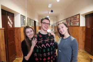 Trójka uczniów na szkolnym korytarzu, uśmiechnięci, pozują do zdjęcia (jeden w świątecznym swetrze).