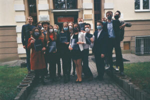 Grupa uczniów przed budynkiem szkoły, większość w maseczkach, trzymają teczki/świadectwa.