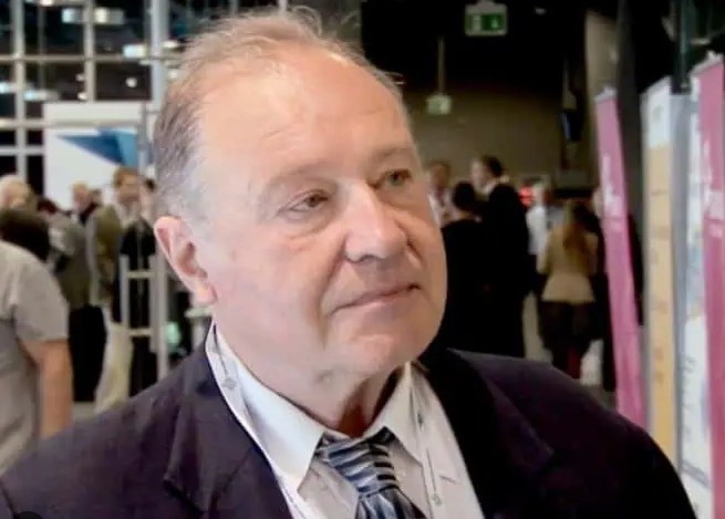 Na zdjęciu widoczny jest portret starszego mężczyzny przedstawionego od klatki piersiowej w górę. Mężczyzna znajduje się na pierwszym planie, lekko zwrócony w prawą stronę kadru, z głową delikatnie przechyloną i wzrokiem skierowanym w bok, jakby słuchał rozmówcy znajdującego się poza kadrem. Jego wyraz twarzy jest spokojny, skupiony i neutralny.Ma krótkie, przerzedzone, siwiejące włosy, zaczesane do tyłu. Czoło jest wysokie, twarz owalna, skóra jasna z widocznymi zmarszczkami charakterystycznymi dla osoby w starszym wieku. Brwi są jasne, oczy średniej wielkości. Nos prosty, usta zamknięte, lekko zaciśnięte w naturalnym, neutralnym ułożeniu.Ubrany jest formalnie: ma na sobie jasną koszulę (najprawdopodobniej białą lub bardzo jasnoniebieską), krawat w odcieniach szarości z ukośnymi, naprzemiennymi paskami oraz ciemną marynarkę – prawdopodobnie granatową lub czarną. Strój sugeruje oficjalne wydarzenie, takie jak konferencja, spotkanie branżowe lub uroczystość.W tle, które jest lekko rozmyte, widać wnętrze dużej sali. Znajdują się tam inne osoby – kilka postaci stojących i rozmawiających w grupach. Po prawej stronie kadru widoczna jest pionowa plansza lub roll-up w jasnym kolorze (różowym lub pastelowym), co dodatkowo wskazuje na wydarzenie targowe, konferencyjne lub wystawowe. W górnej części tła można dostrzec elementy konstrukcyjne budynku – metalowe ramy, przeszklone fragmenty ścian oraz oświetlenie sufitowe.Całość sprawia wrażenie ujęcia wykonanego podczas wywiadu lub rozmowy w trakcie wydarzenia publicznego. Zdjęcie ma charakter reportażowy, skupiający uwagę na twarzy i wyrazie osoby znajdującej się na pierwszym planie.