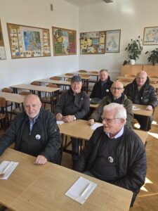 Sala lekcyjna z rzędami drewnianych ławek i krzeseł. Przy pierwszych ławkach siedzi sześciu starszych mężczyzn w kurtkach, część z przypinkami; na stołach leżą kartki i długopisy. Na ścianach wiszą tablice z mapami i materiałami edukacyjnymi. Wnętrze jasne, oświetlone światłem dziennym.