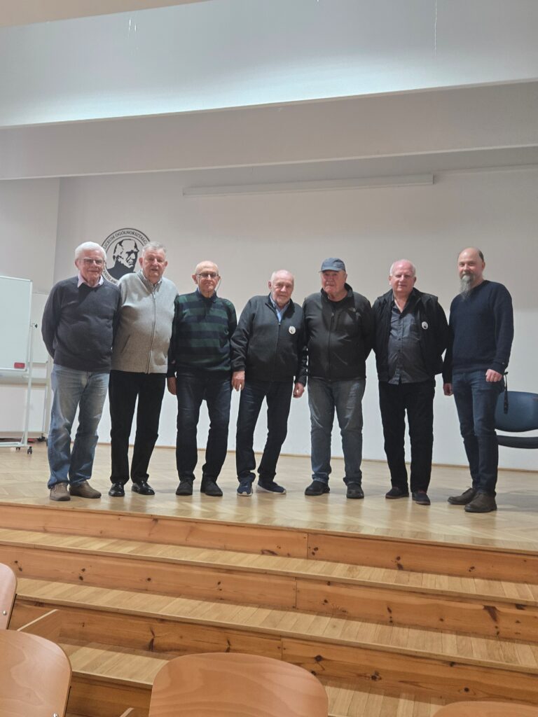 Sześciu starszych mężczyzn stoi w rzędzie na drewnianej scenie w sali szkolnej. Za nimi jasna ściana z godłem lub logo szkoły. Ubrani są w swetry i kurtki, stoją wyprostowani, zwróceni w stronę aparatu.