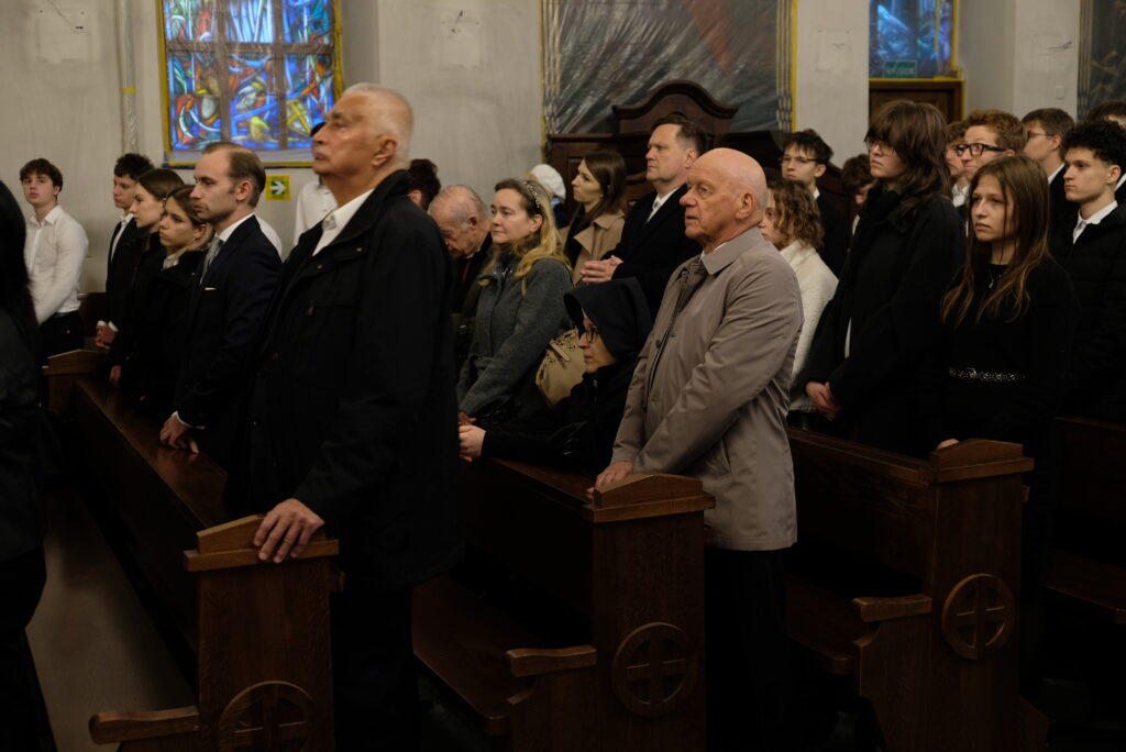 Zaproszeni goście stoją w kościelnych ławkach.
