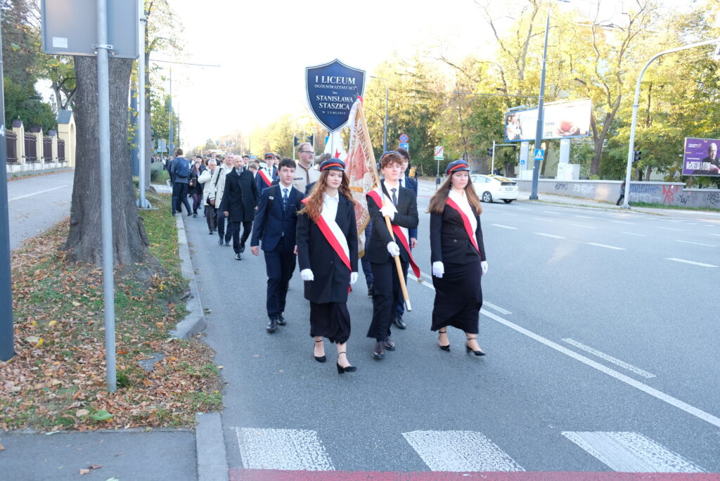 Uczniowie z biało-czerwonymi szarfami maszerują ze sztandarem.