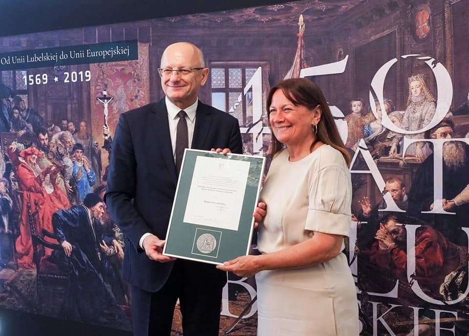 Mężczyzna w ciemnym garniturze i kobieta w jasnej sukience wspólnie prezentują oprawiony dokument podczas oficjalnej uroczystości. W tle znajduje się dekoracyjna plansza z historycznymi obrazami i datami „1569–2019”.