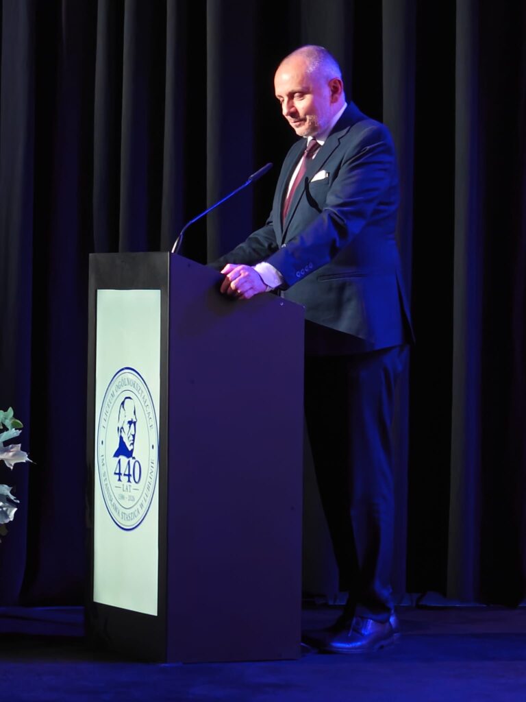 Mężczyzna przemawia przy mównicy z jubileuszowym emblematem.