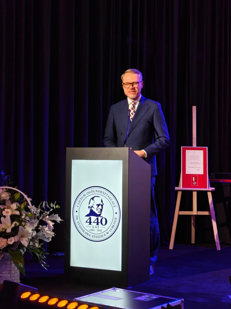 Mężczyzna przemawia przy mównicy z jubileuszowym emblematem.
