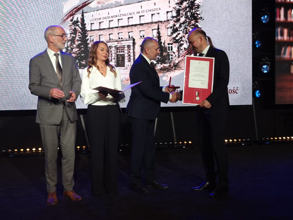 Mężczyzna prezentuje oprawione wyróżnienie, obok stoi kobieta w białej marynarce.