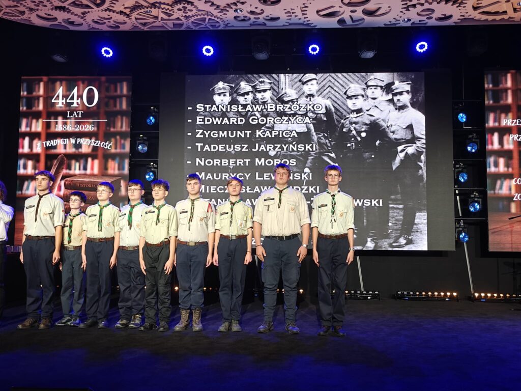 Grupa młodych wykonawców w jasnych koszulach stoi w szeregu na scenie.