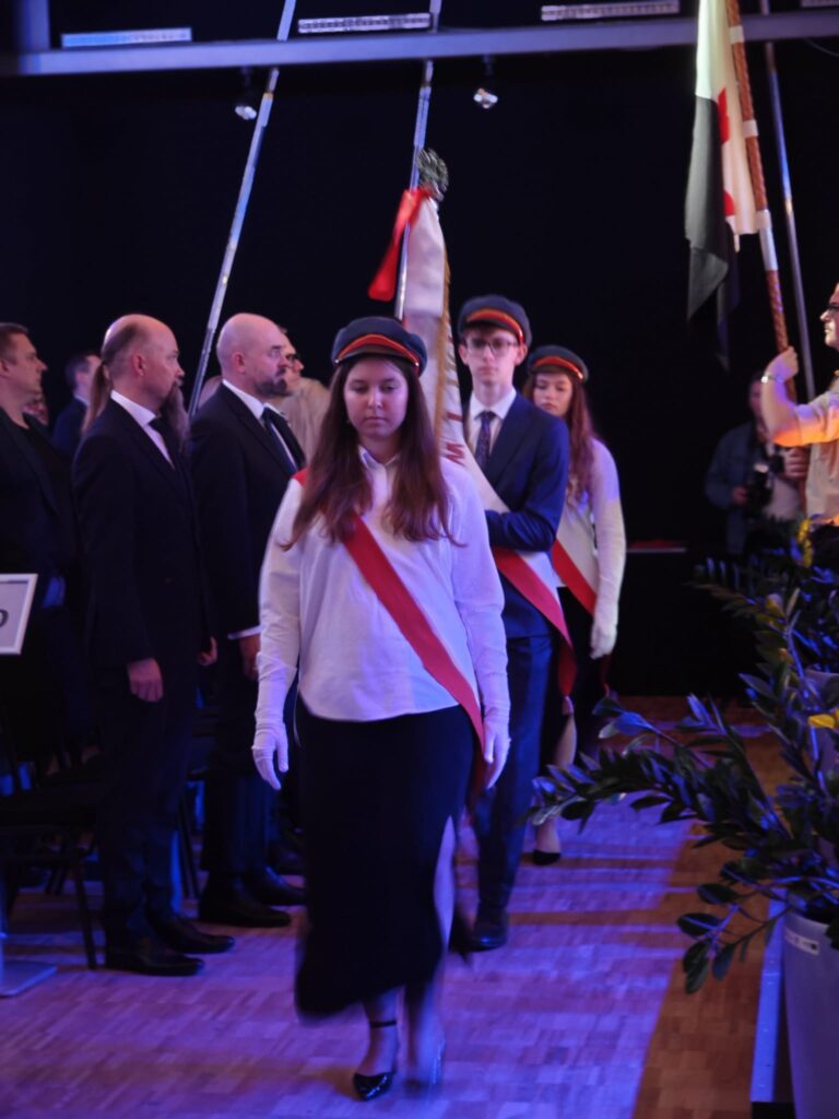 Poczet sztandarowy w biało-czerwonych szarfach wchodzi do sali widowiskowej.
