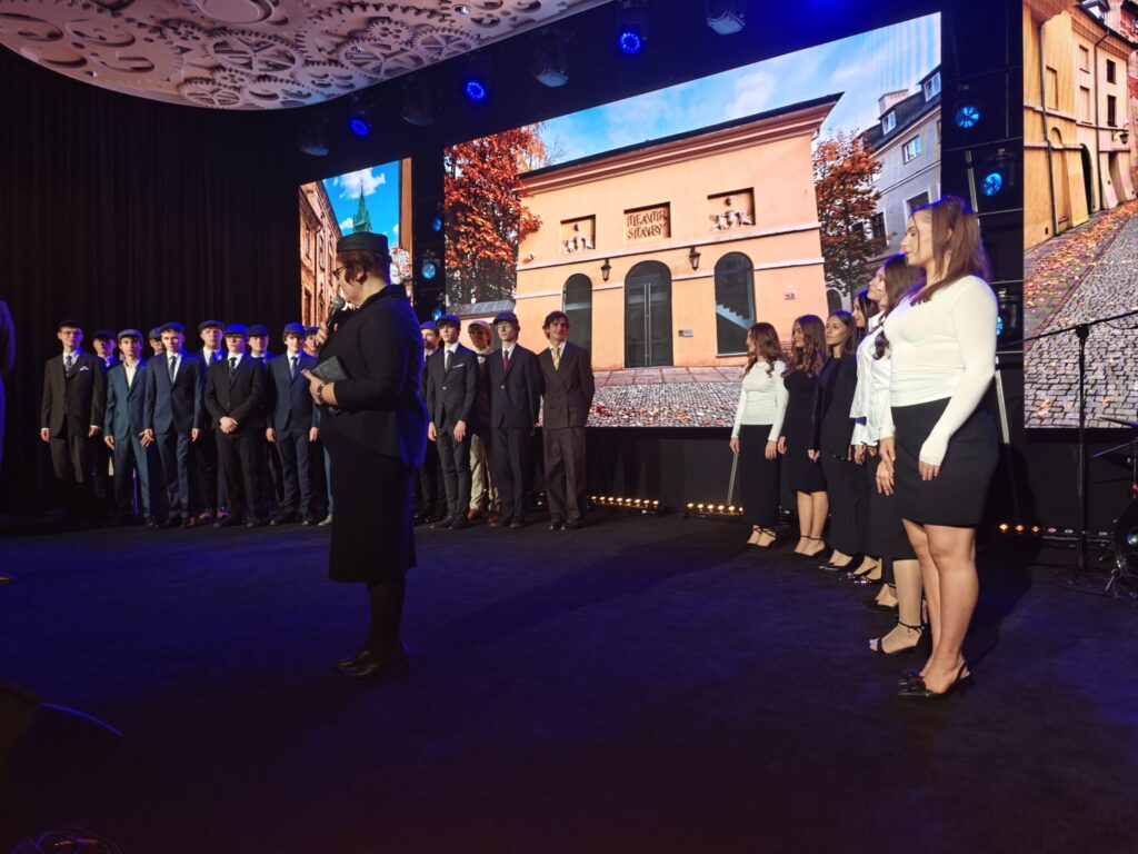 Kobieta przemawia na scenie, za nią stoi grupa młodzieży w eleganckich strojach.