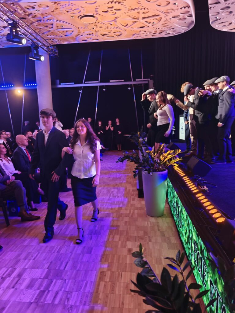 Młodzi uczestnicy uroczystości przechodzą wzdłuż sceny przy kolorowym oświetleniu.