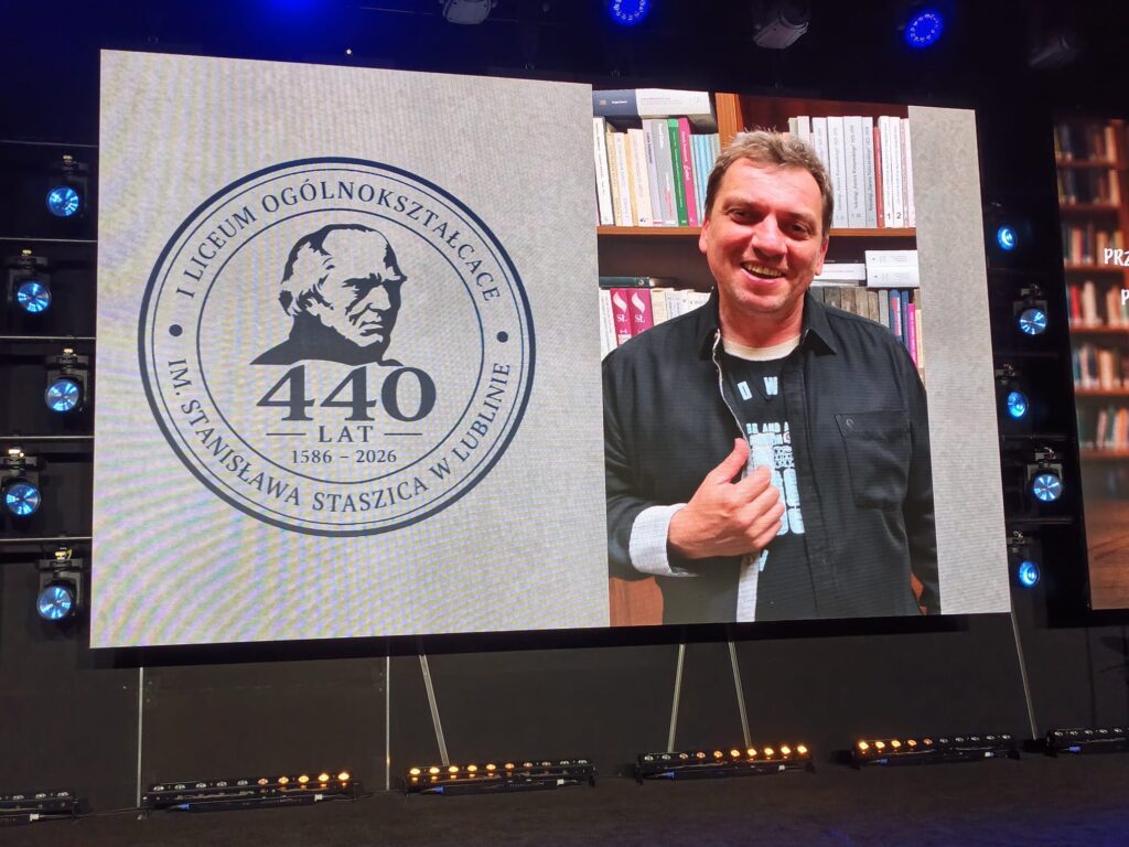 Uśmiechnięty mężczyzna pokazuje uniesiony kciuk na tle emblematu 440-lecia szkoły.