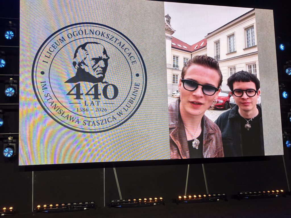 Dwie osoby pozują do selfie przed jubileuszowym emblematem szkoły.