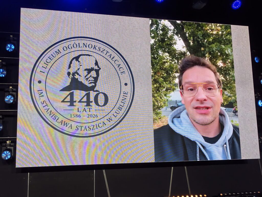 Mężczyzna w jasnoniebieskiej koszuli widoczny na ekranie obok emblematu 440-lecia.