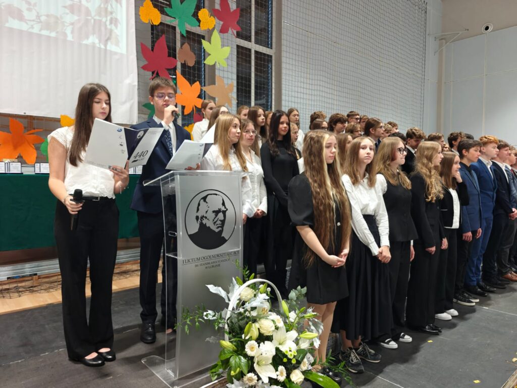 Uczennica przemawia przy mównicy, za nią stoi grupa młodzieży.