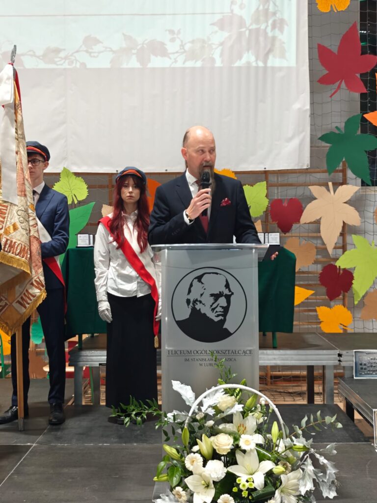 Mężczyzna przemawia przy mównicy podczas uroczystości szkolnej, obok stoi poczet sztandarowy.