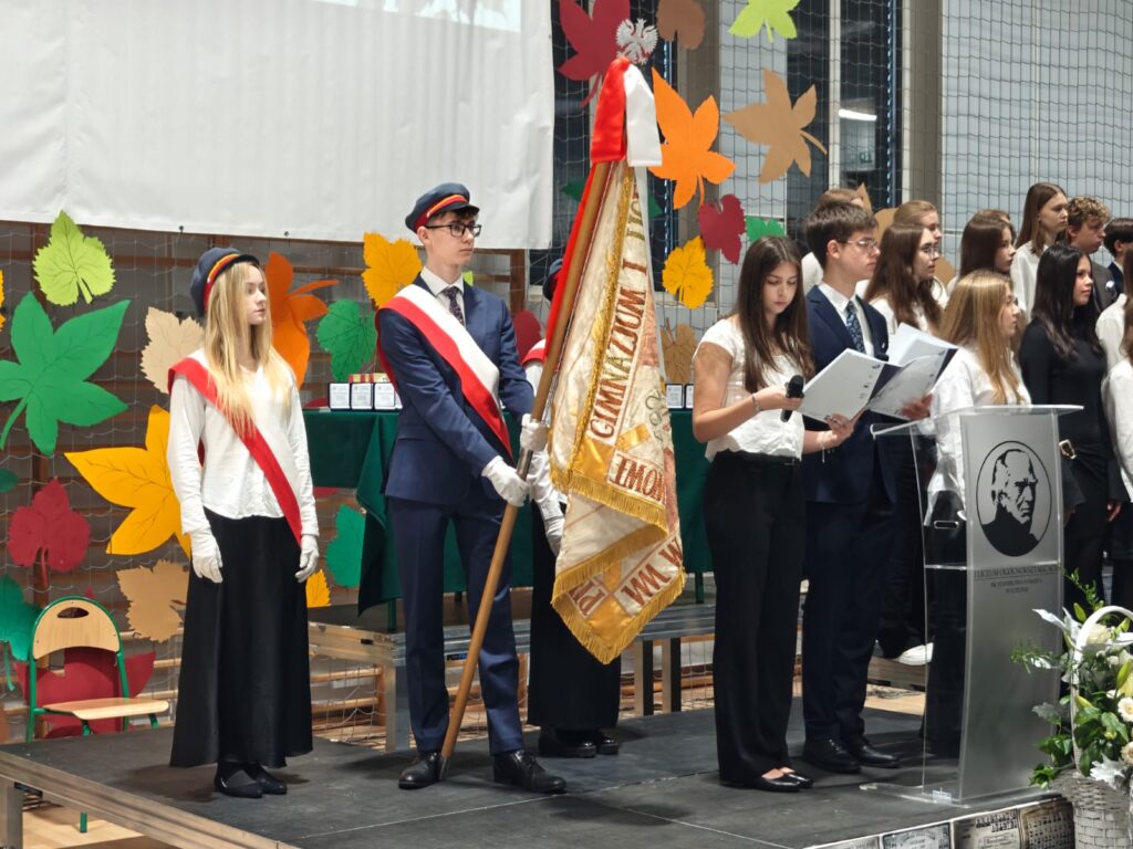 Poczet sztandarowy stoi obok uczniów podczas szkolnej ceremonii.