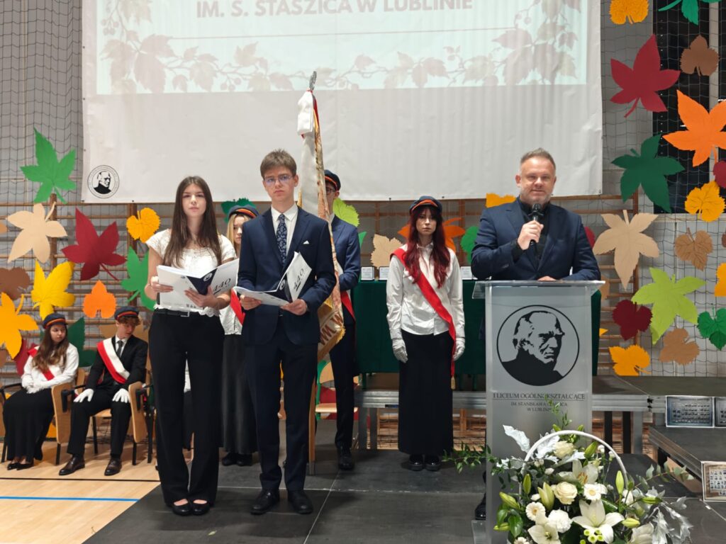 Grupa uczniów z dokumentami i poczet sztandarowy stoją przy mównicy.